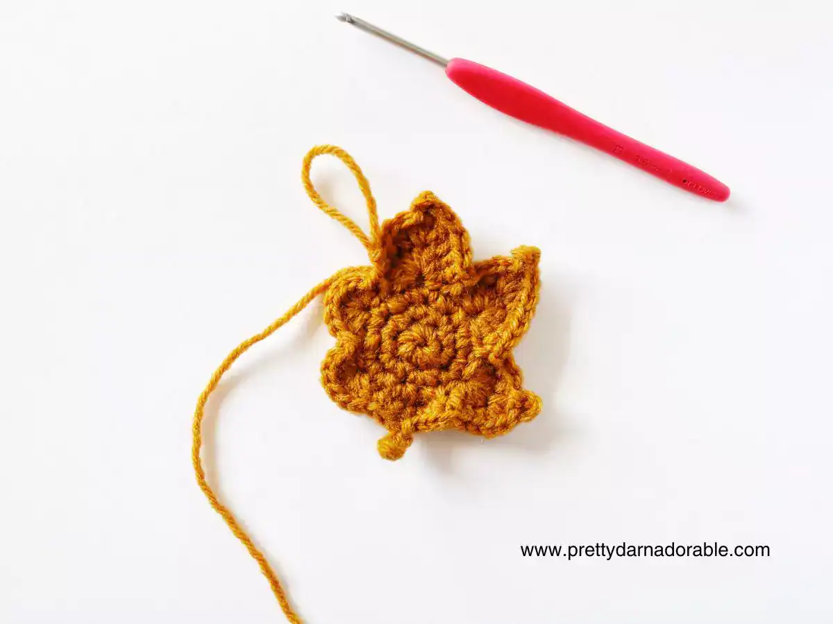 A mustard yellow crocheted maple leaf with a loose yarn end lies on white beside a red-handled crochet hook; www.prettydarnadorable.com. Free crochet pattern