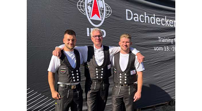 Ein Mann in traditioneller Zunftkleidung steht vor einem schwarzen Banner mit weiß-rotem Emblem und lächelt in die Kamera.