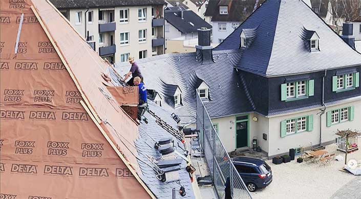 Dächer mit grauen Schindeln und kleinen Fenstern, einige Arbeiter auf dem Dach, umliegende Wohngebäude im Hintergrund.