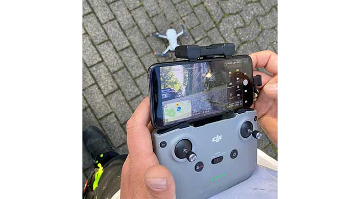 Eine Hand hält eine Drohnenfernbedienung mit Smartphone-Display, das die Kamerasicht und eine Karte zeigt; im Hintergrund liegt eine kleine Drohne auf Pflastersteinen.