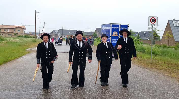 Zwei Männer in traditioneller schwarzer Tracht und Zylinderhut gehen auf einer Straße, einer hält einen Stock. Im Hintergrund stehen weitere Menschen.