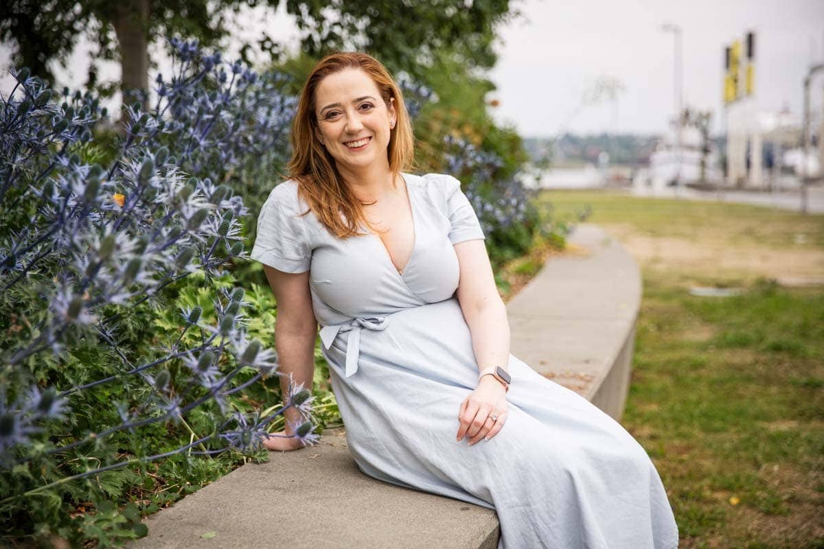 A woman in a light blue dress sits on a concrete ledge by purple flowers, smiling. The background includes grass, trees, and a blurry view of a waterfront.
