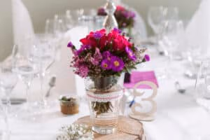 Hochzeitslocation Haus Runde 7 Eine Glasvase mit rosa und violetten Blumen schmückt einen Hochzeitstisch, neben dem Tafelaufsatz mit der Zahl 3.