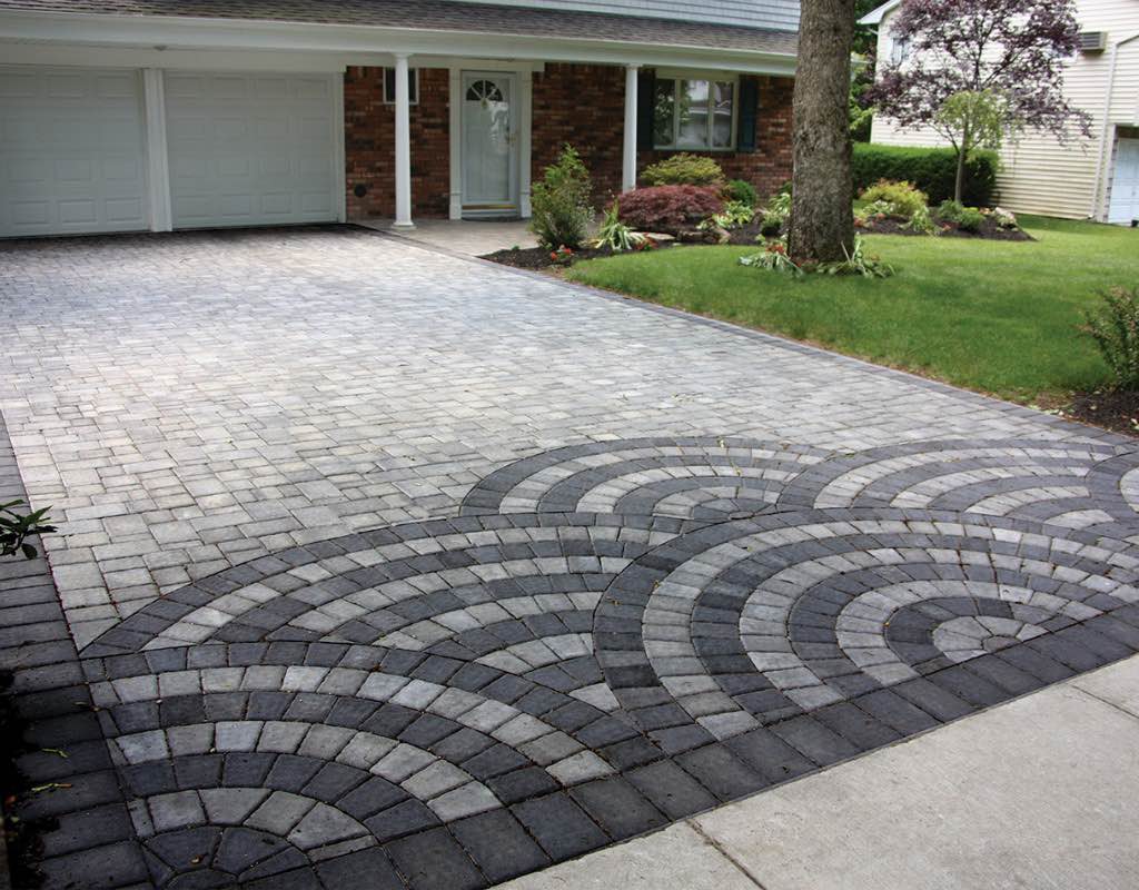 A brick driveway with a circular pattern leads to a double garage attached to a single-story brick house with a manicured lawn.