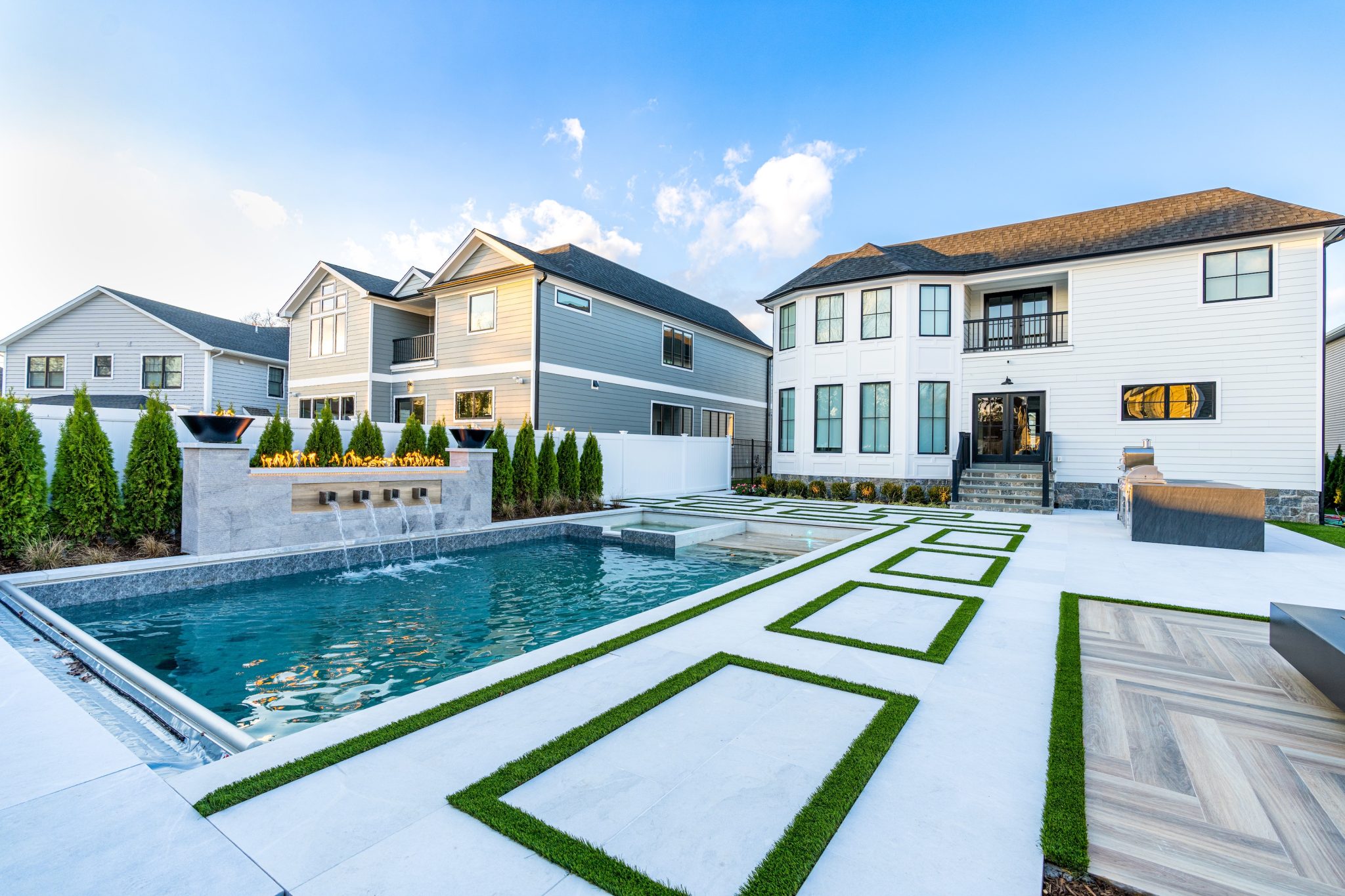 Modern backyard with a rectangular swimming pool, geometric stone and grass patio, fire feature, and a white two-story house in the background.