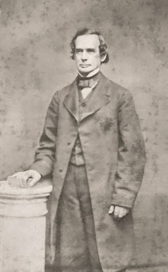 A black and white historic photo of a 19th-century man in formal attire standing beside a pedestal.