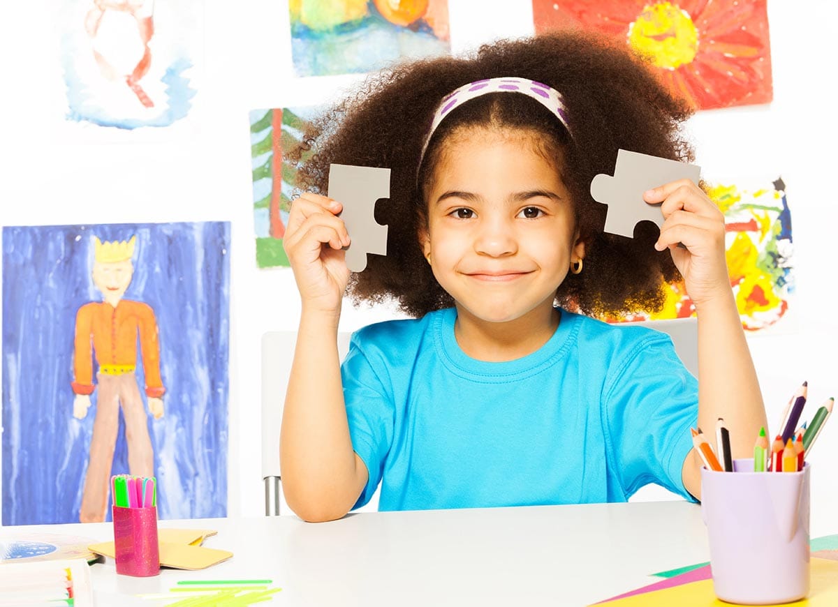 A young girl with curly hair holds two large puzzle pieces and smiles, sitting at a table with art supplies. Colorful children's artwork is displayed on the wall behind her.
