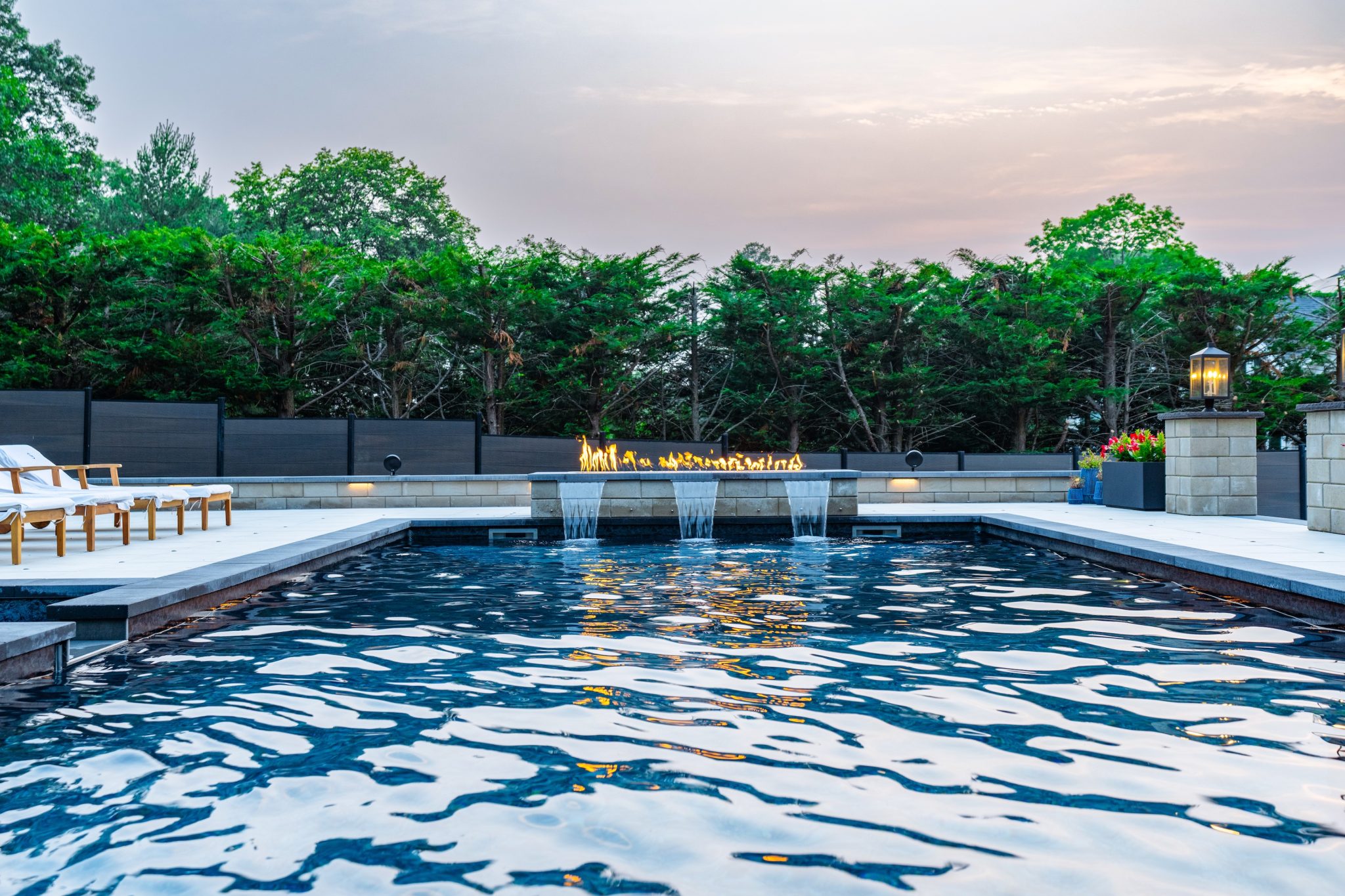 Rectangular outdoor pool with three water features, surrounded by lounge chairs, fencing, greenery, and a lit fire feature at dusk.