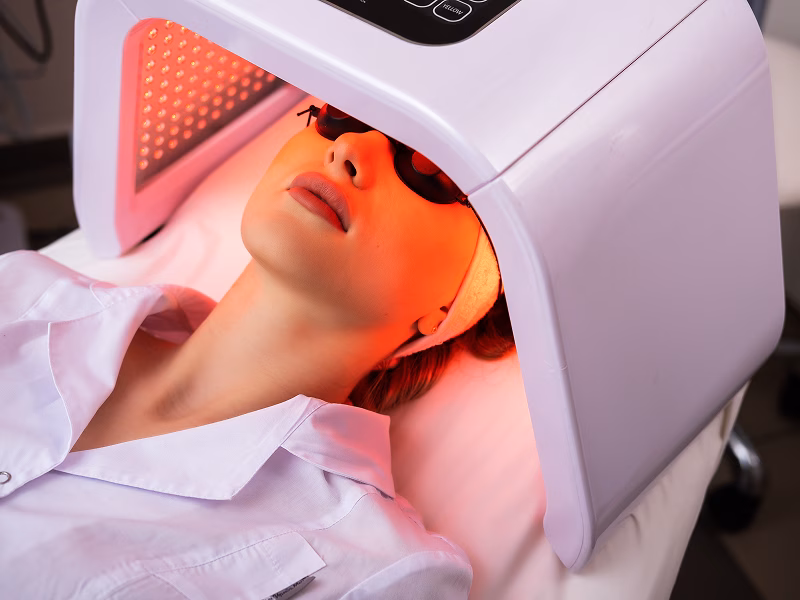 Person liegt mit Brille unter einem kosmetischen LED-Lichtgerät in einem Behandlungsraum