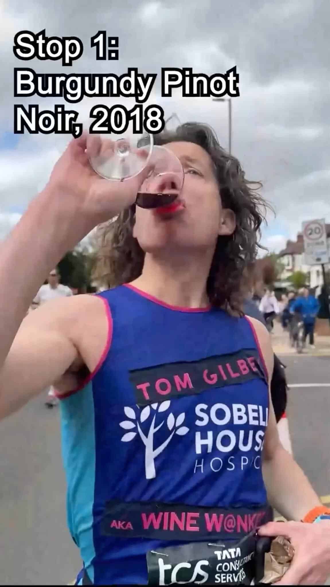 A marathon runner, Tom Gilbey, drinks wine mid-race during the London Marathon's wine tasting challenge. The text on his vest reads "Tom Gilbey" and "Sobell House Hospice." He's participating in a charity run, part of a viral TikTok video.