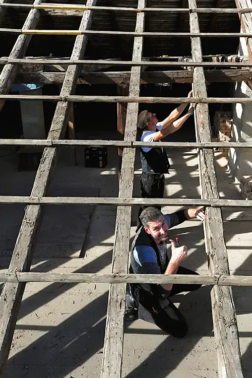 Zwei Männer arbeiten unter Holzbalken eines teilweise abgedeckten Daches in einer Baustellenumgebung, Sonnenlicht fällt auf die Szene.