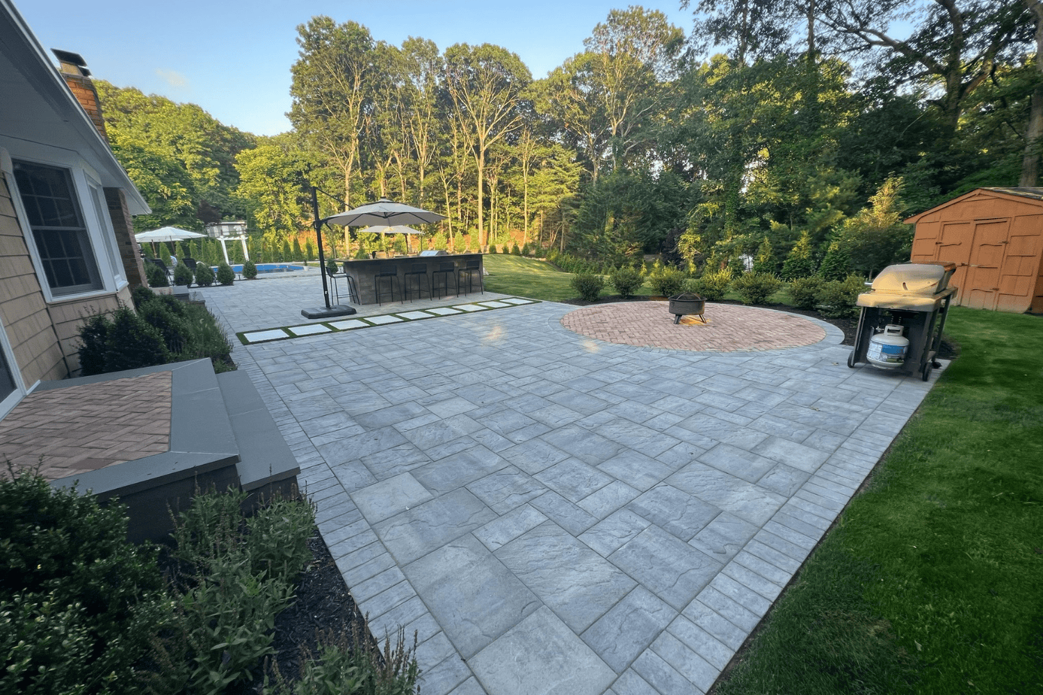 Spacious backyard patios feature gray stone pavers, a round fire pit area, a grill, outdoor seating with umbrella, and a pool in the background, all surrounded by lush trees and greenery.