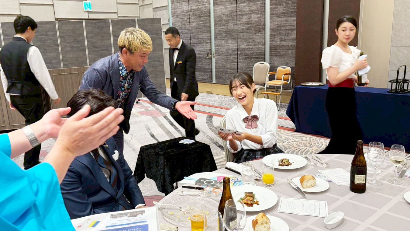 金沢ロータリークラブ パーティーでのマジックショー|石川・富山の笑顔を広げる活動報告 2 金沢ロータリークラブのパーティーで、マジシャンがテーブルマジックを披露し、観客がカードを手に笑顔で楽しんでいる様子。