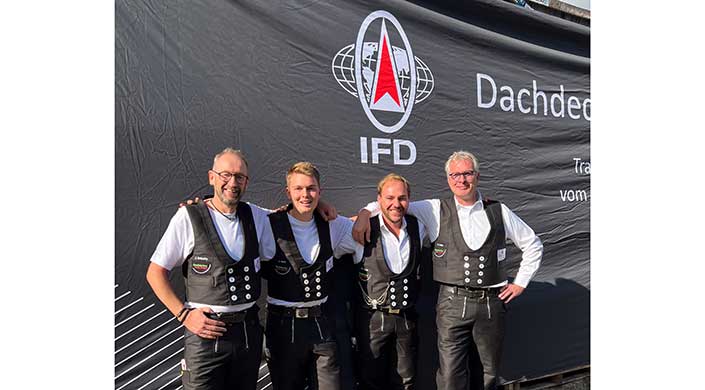 Zwei Männer in traditioneller Arbeitskleidung stehen vor einem Banner mit dem IFD-Logo und einer Weltkugel-Grafik, lächeln und posieren gemeinsam für ein Foto.