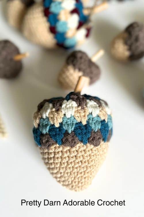 Close-up of a blue, white, beige, and brown crocheted acorn among others on white. Text: Pretty Darn Adorable Crochet. Free crochet pattern