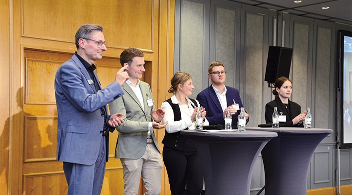 Drei junge Menschen in formeller Kleidung stehen an einem Stehtisch, unterhalten sich und applaudieren in einem hellen Konferenzraum.