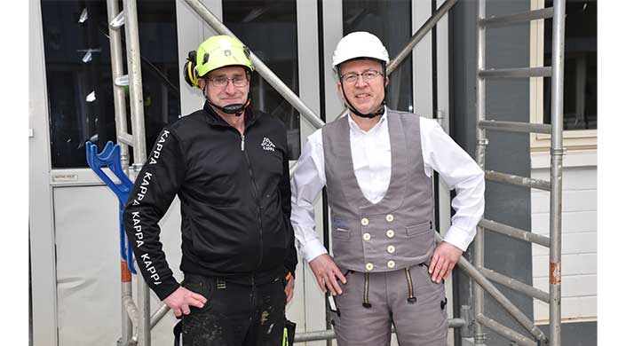 Zwei Männer mit Schutzhelmen stehen vor einem Gebäude; einer trägt Arbeitskleidung, der andere eine weiße Weste mit Hemd und Brille.