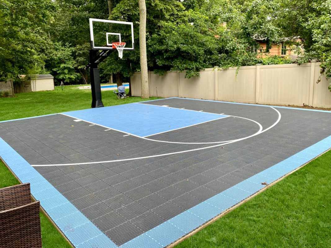 Outdoor residential basketball court with a blue and black surface, backboard, and surrounding greenery. Perfect for those seeking a quality sports court backyard installation in Huntington.