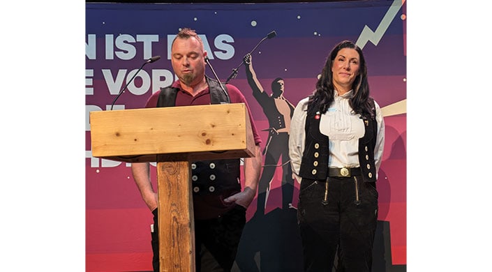Bild von Rede von Lehrlingswart für seine Azubis Ein Mann steht an einem Rednerpult, im Hintergrund ist eine Illustration einer historischen Figur mit erhobenem Arm und dunklem Himmel zu sehen.