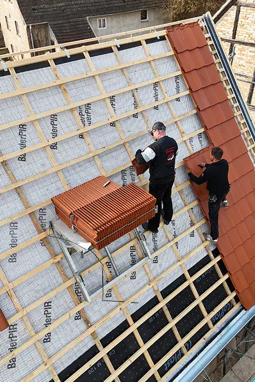 Ein Arbeiter auf einem Dach installiert Dachziegel, während er auf einer Holzkonstruktion steht. Stapel mit roten Dachziegeln sind neben ihm platziert.