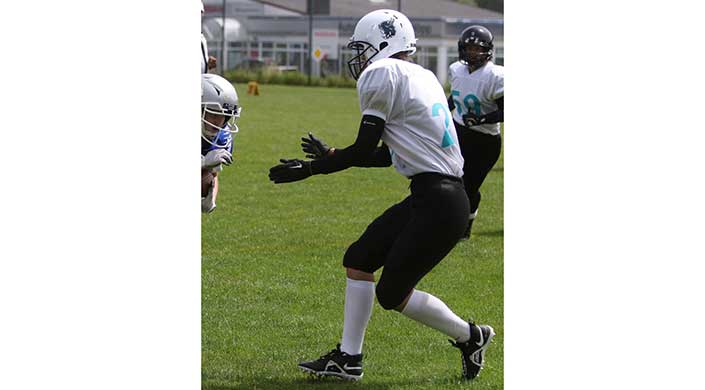 Ein American-Football-Spieler im weißen Trikot und Helm steht auf einem grünen Spielfeld, bereit, den Ball zu fangen oder einen Spielzug auszuführen.