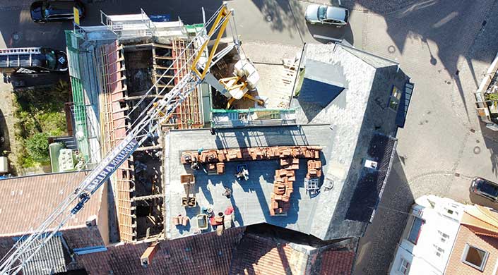 Luftaufnahme einer Baustelle mit Bauarbeitern, gestapelten Ziegeln und einem Kran, der Material auf das Dach hebt.