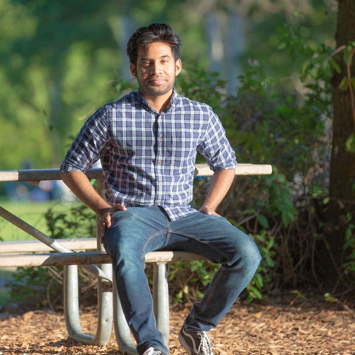 A man in a plaid shirt and jeans sits on a picnic table bench in a park, surrounded by greenery and sunlight.