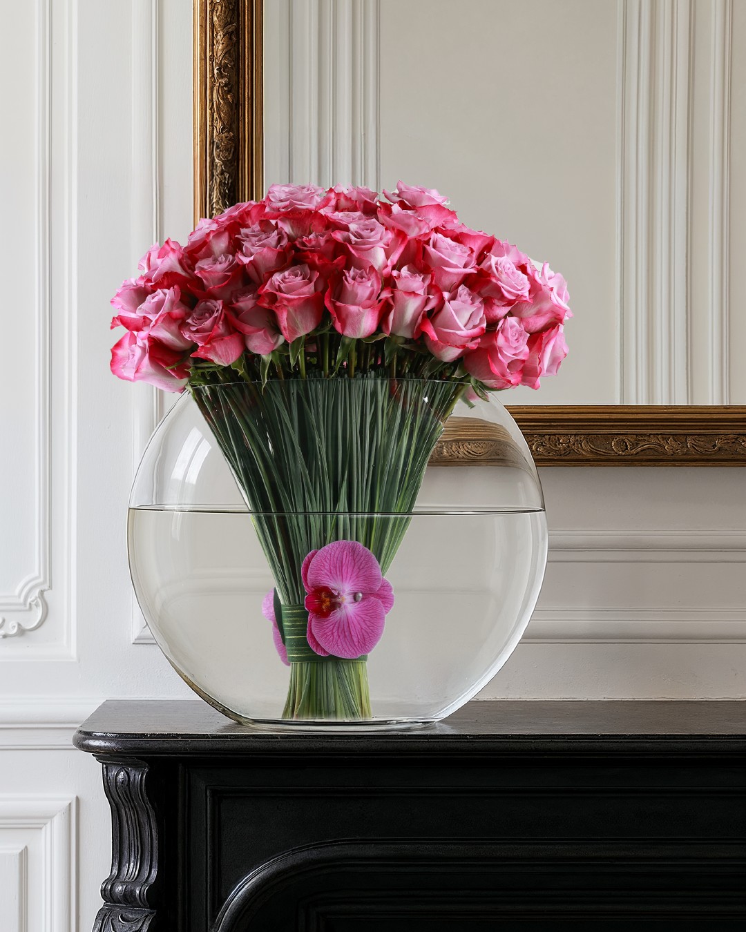 A dramatic French Florist arrangement of pink roses and a floating orchid in a large round glass bowl vase displayed on a dark mantle.