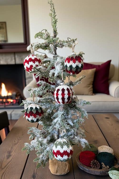 A small, snow-dusted Christmas tree with red, white, and green yarn ornaments sits on a wooden table; yarn and pinecones nearby. Free easy crochet Christmas Bauble pattern