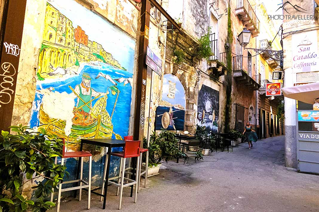 Stühle einer Bar stehen vor bunten Wandgemälden der engen Altstadtgassen von Taranto