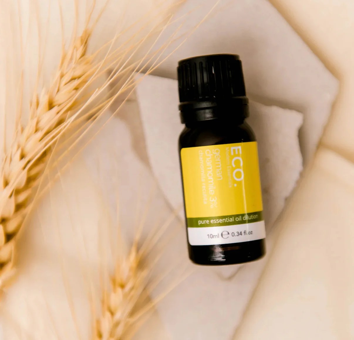 A 10ml bottle of German Chamomile 3% in Grapeseed Oil sits on a white stone, encircled by wheat stalks against a beige backdrop.