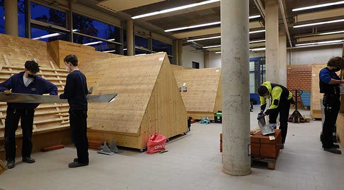 Holzverschläge mit dreieckiger Form in einem Innenraum, sichtbare Elektrik an den Wänden, ein roter Beutel auf dem Boden vorne.