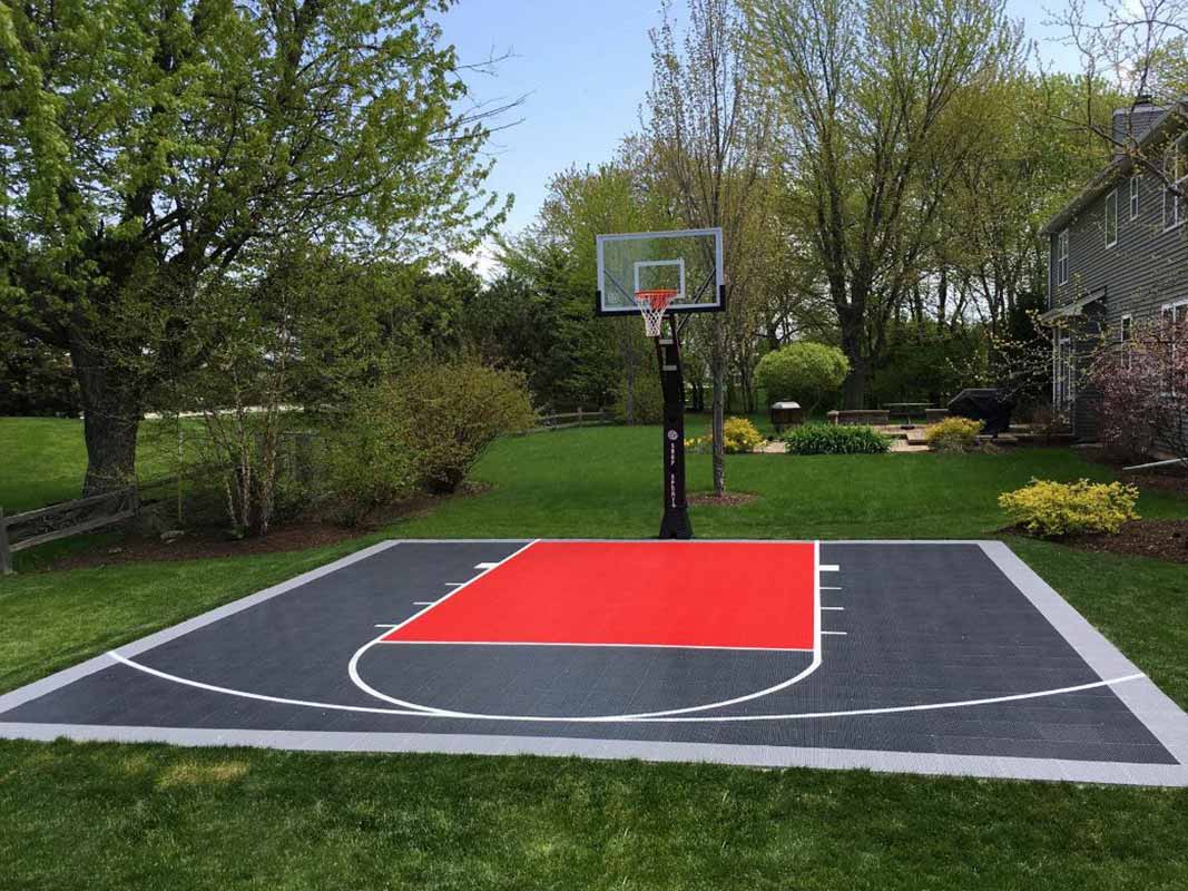Outdoor basketball half-court with a red key area, surrounded by grass and trees, featuring a house in the background – perfect for sports court backyard installation in Huntington.