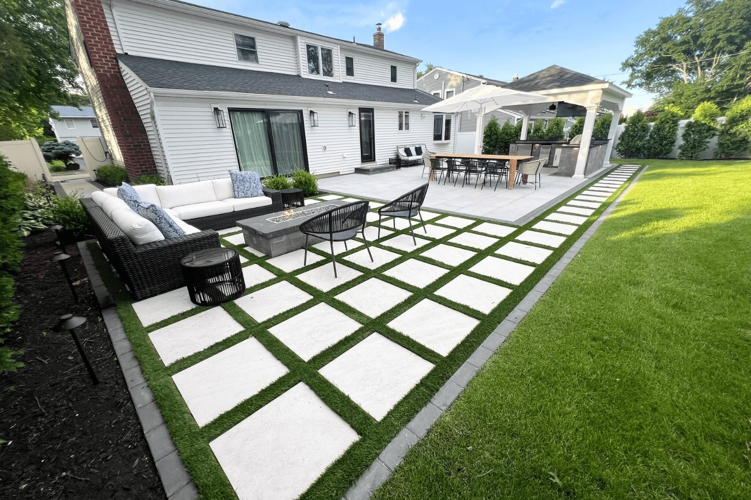 Modern backyard patio with white stone pavers set in grass, a stylish outdoor seating area, cozy fire pit, bar with stools, and covered pergola—showcasing how patios can elevate the charm of a lawn behind a white two-story house.