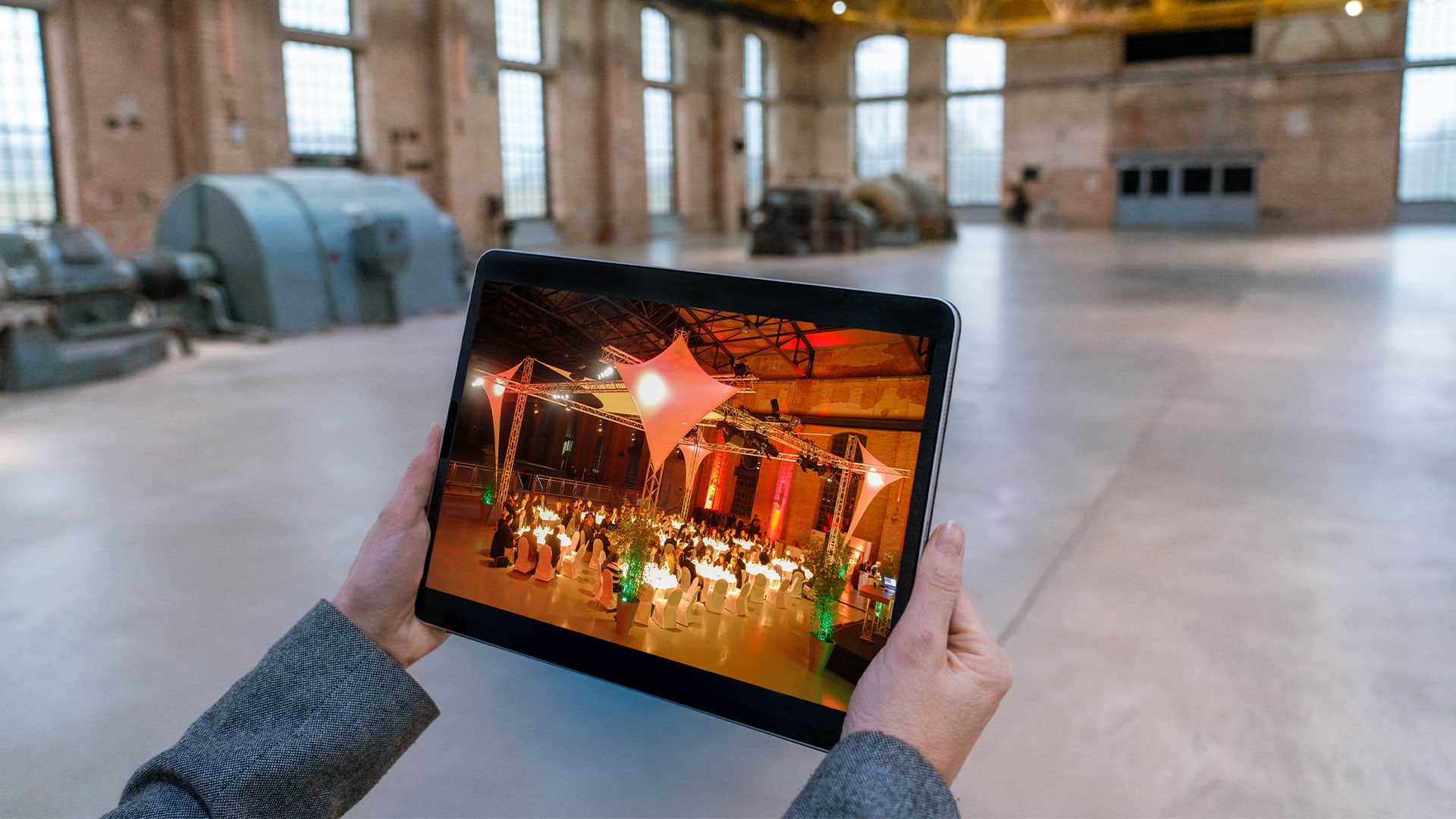 Ein Foto von einem Tablet das von einer Person gehalten wird. Auf dem Tablet ist eine belebte Abendveranstaltung in der Halle zu sehen.