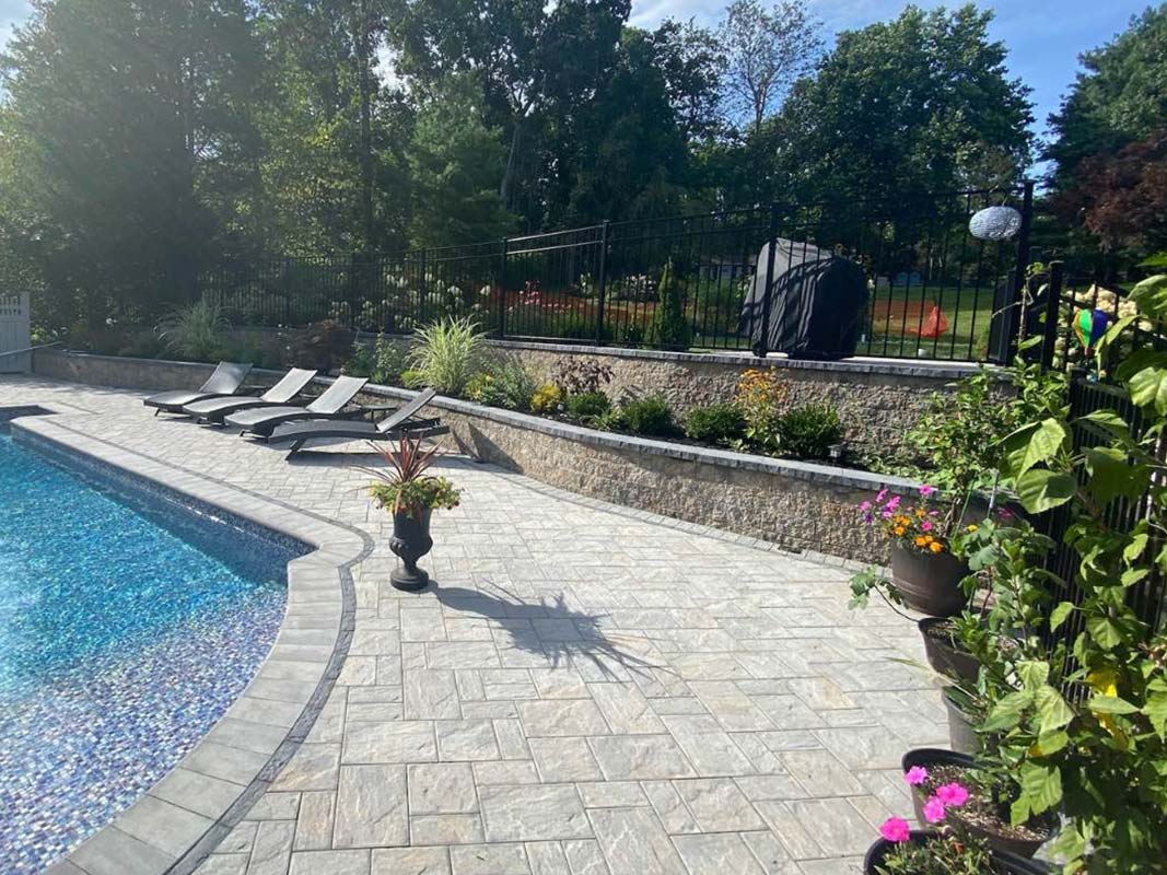 A backyard pool area with four lounge chairs, various potted plants, a built-in stone grill, and trees in the background. A terrace retaining all borders the pool area with flower plantings in Elwood.