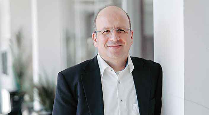 Ein Mann mit Brille und Glatze trägt ein weißes Hemd und schwarzes Sakko, lächelt leicht, unscharfer Hintergrund.