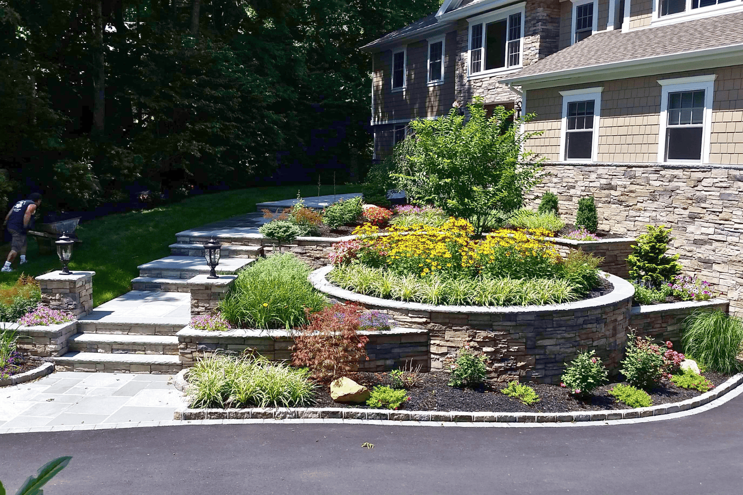 A beautifully crafted landscape design features stone retaining walls, tiered garden beds, a small tree, flowering plants, and a stone staircase leading to a house with tan siding and elegant stone accents.