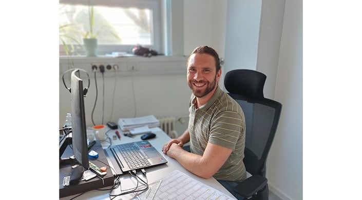 Bild von Lehrlingswart Geschke an seinem Schreibtisch Ein lächelnder Mann sitzt an einem Schreibtisch mit Laptop, Papieren und Kalender in einem hellen Büro.