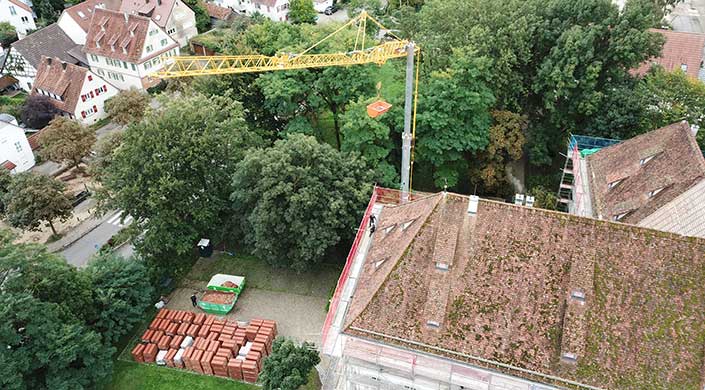 Blick von oben auf einen Baukran, der Baumaterial auf ein Gebäudedach hebt, umgeben von Bäumen und Gerüst.