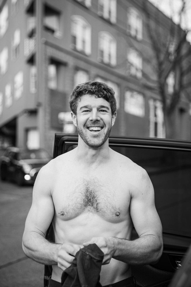 Man standing shirtless by a car door on a street, smiling at the camera. Background shows buildings and a tree. Black and white image.