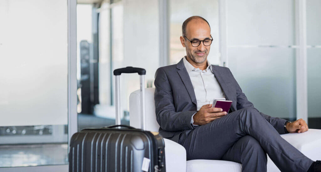 Geschäftsmann mit Koffer am Flughafen nutzt Smartphone als Symbol für internationale Steuerresidenz und UAE Golden Visa Geschäftsmann mit Koffer am Flughafen nutzt Smartphone als Symbol für internationale Steuerresidenz und UAE Golden Visa