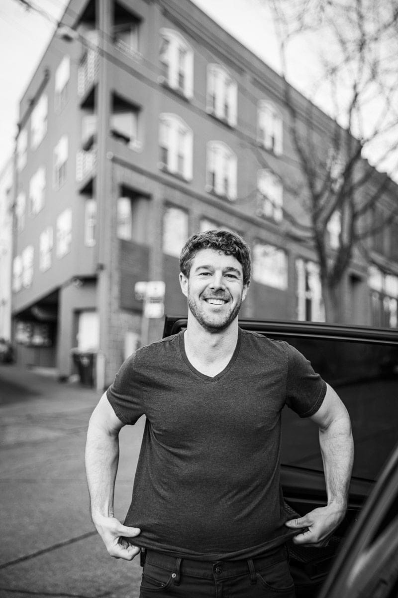 Man smiling while standing by a car, adjusting his t-shirt in front of a multi-story brick building in an urban setting. Black and white photo.
