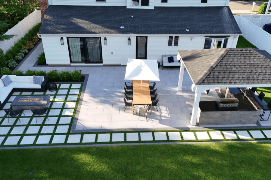 Aerial view of a backyard patio with dining table, chairs under a white umbrella, covered grill area and Outdoor Kitchen, outdoor sofa, fire pit, green lawn bordered by stone pavers—perfect for pavilion installation for ultimate backyard living in Smithtown, NY.