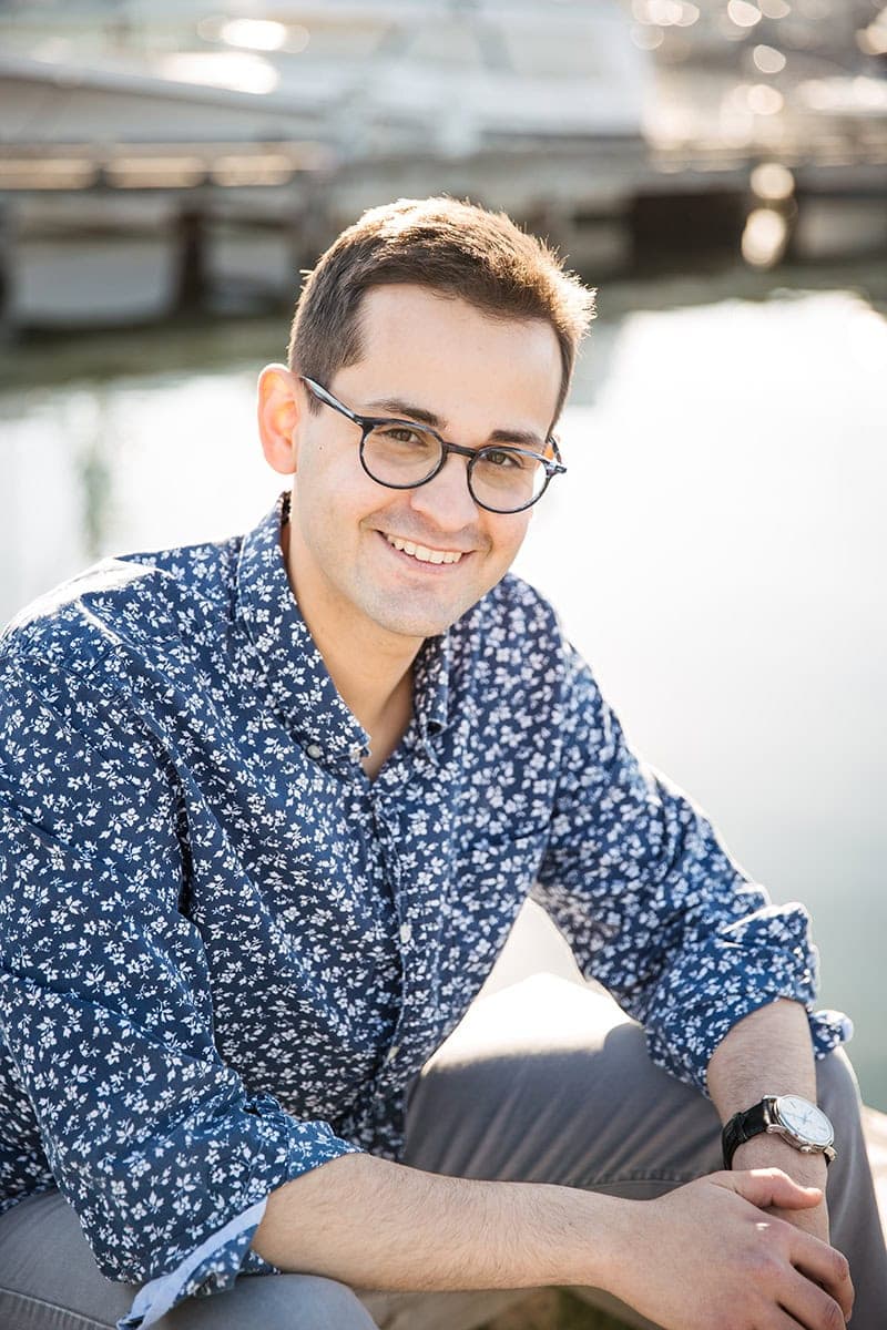 A man wearing glasses and a blue patterned shirt sits outdoors by the water, smiling at the camera with boats and docks in the background.