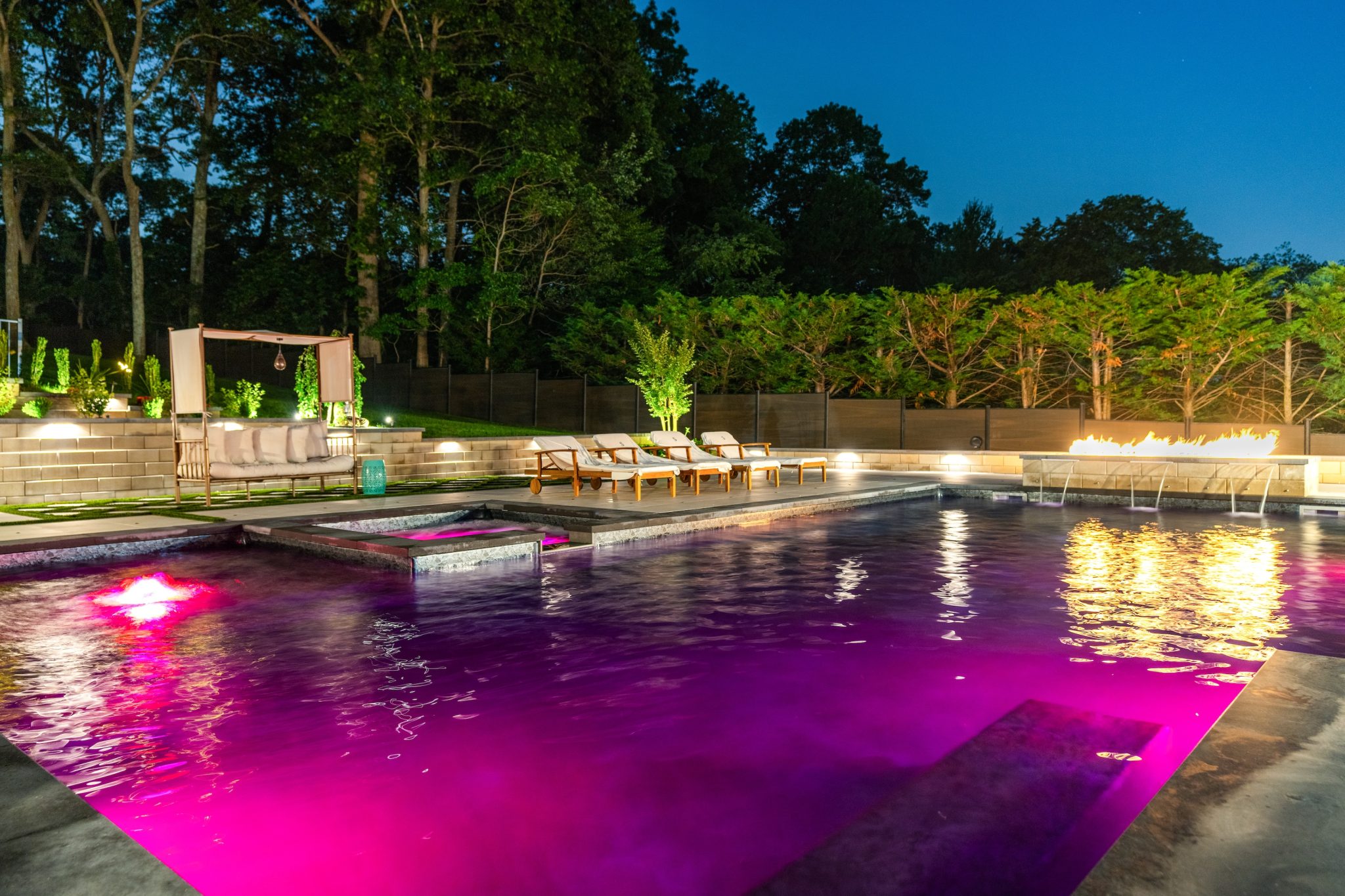 A modern backyard pool at night with purple lighting, lounge chairs, a fire feature, and a canopy seating area, surrounded by trees and shrubs.