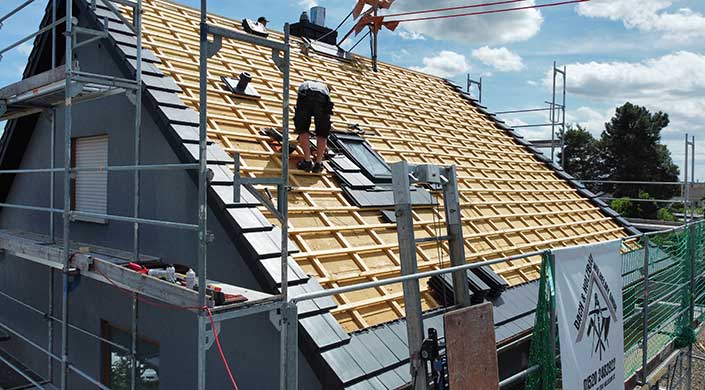 Ein Arbeiter verlegt schwarze Dachziegel auf einem Dach mit Holzlattung an einem sonnigen Tag, Gerüst im Vordergrund.