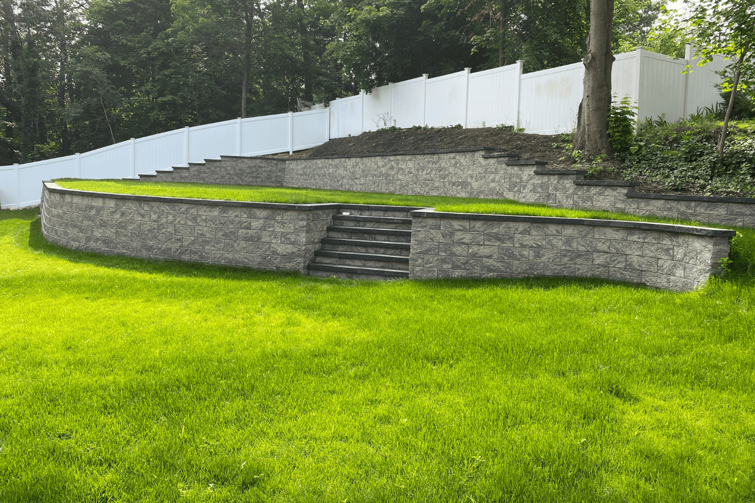Stone retaining walls with steps are built into a grassy yard, creating an elegant landscape feature, with a white fence and trees in the background.