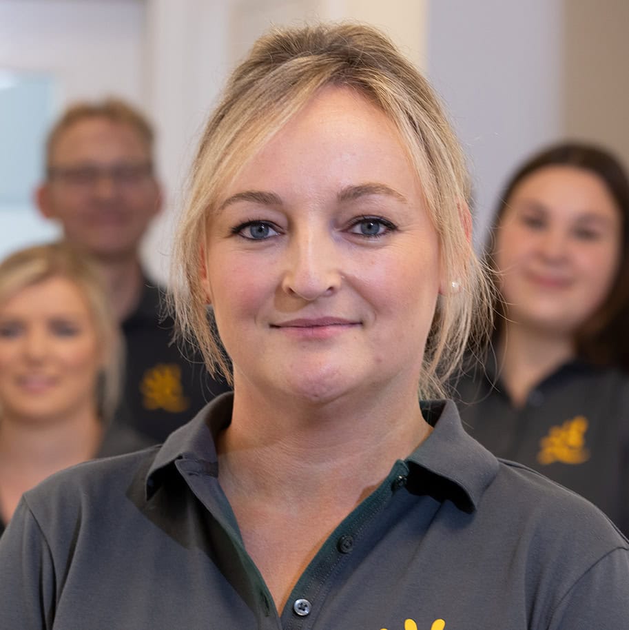 Blonde Frau mit dem grauen Praxis-Poloshirt vor einer Gruppe Mitarbeiter im Hintergrund.