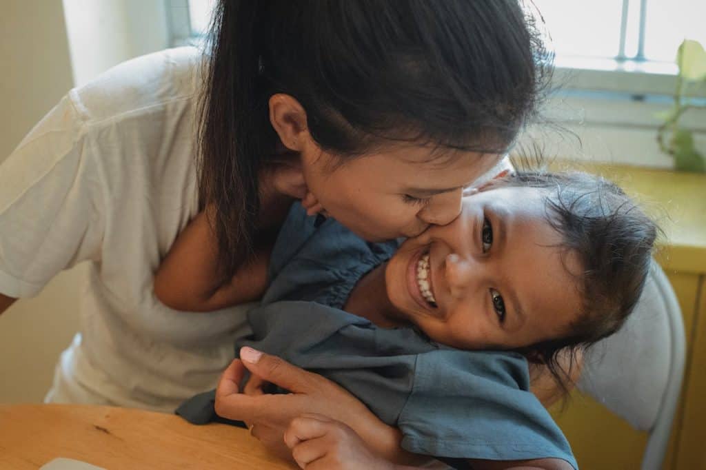 A mother holds her child, kissing her cheek while the child laughs.